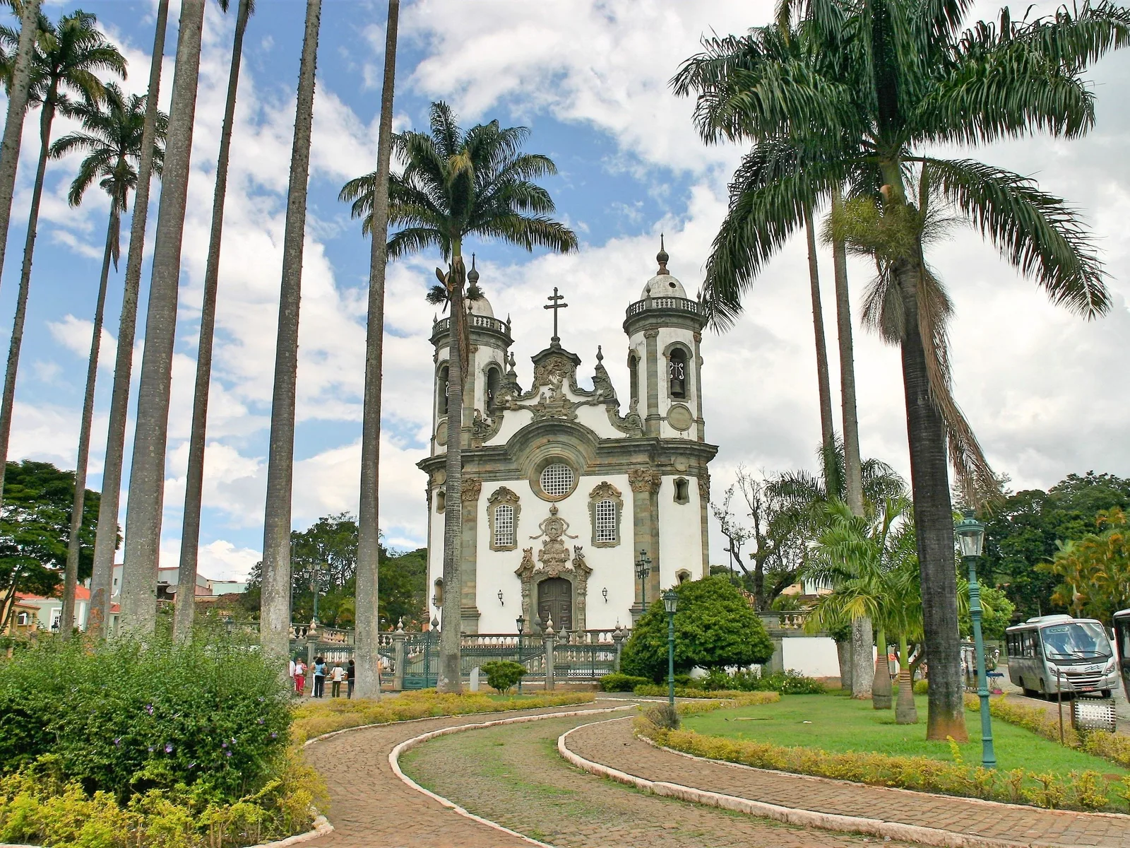 The Igreja de São Francisco de Assis in São João del-Rei seen across its square through tall royal palms, the white church with its rounded corner towers standing behind a low wall and a paved forecourt