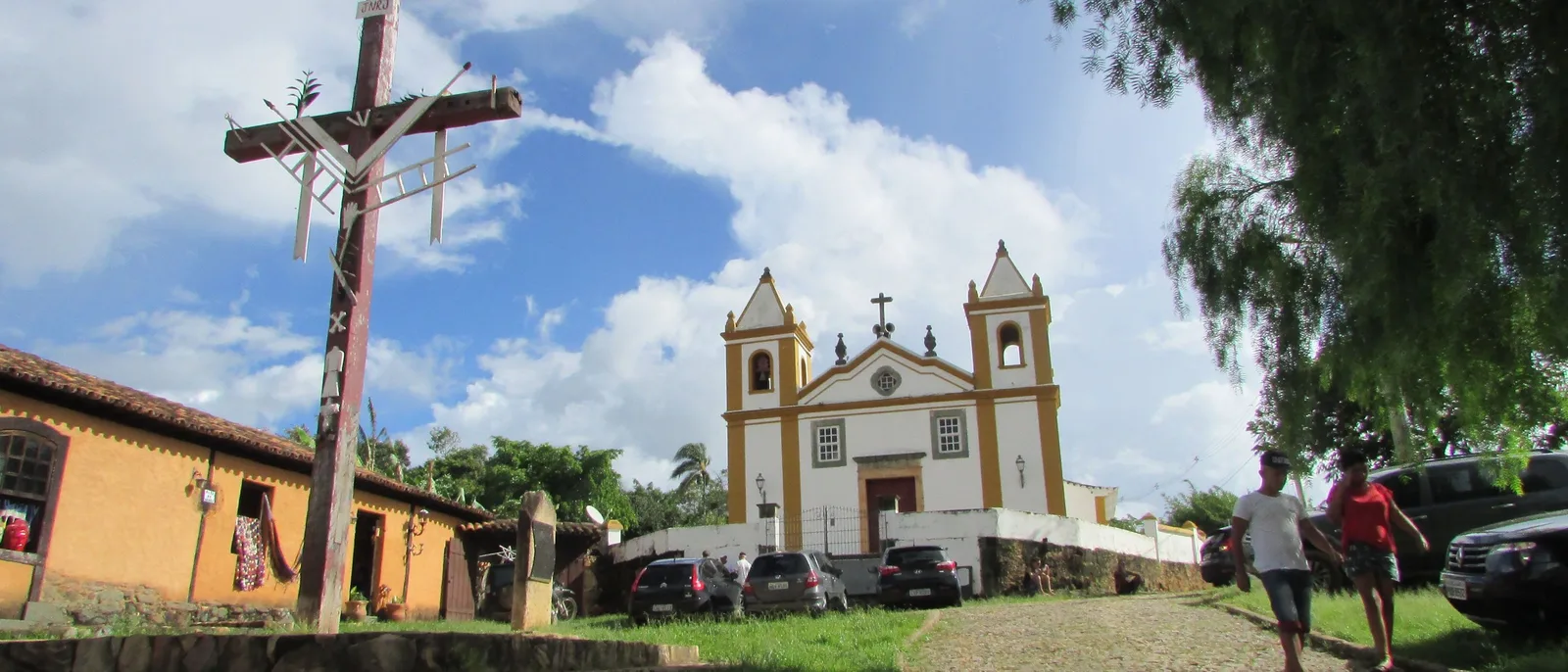 The white-and-yellow twin-towered church at Bichinho above its low wall, a tall weathered timber cruzeiro standing in the foreground, an ochre village house with crafts hung outside it at the left and two people walking up the cobbled slope at the right