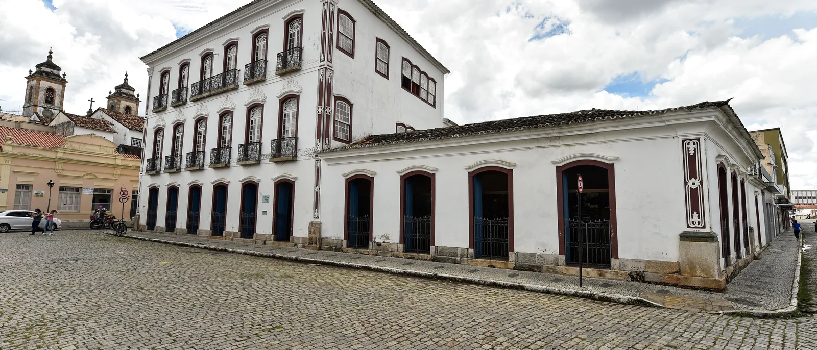 A wide cobbled square in São João del-Rei lined with long white colonial buildings, church towers showing above the rooftops at the far end