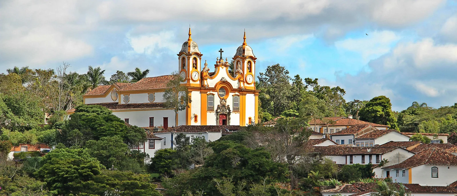 The Matriz de Santo Antônio rising above the trees and red-tiled rooftops of Tiradentes, its yellow-and-white towers catching the light