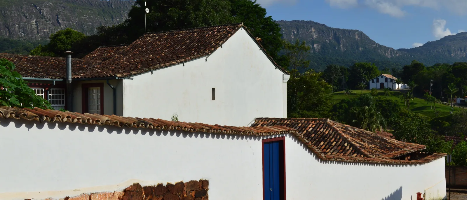 A whitewashed house with a red pantile roof on the upper edge of Tiradentes, with more rooftops below it and the wooded ridge of the Serra de São José filling the background